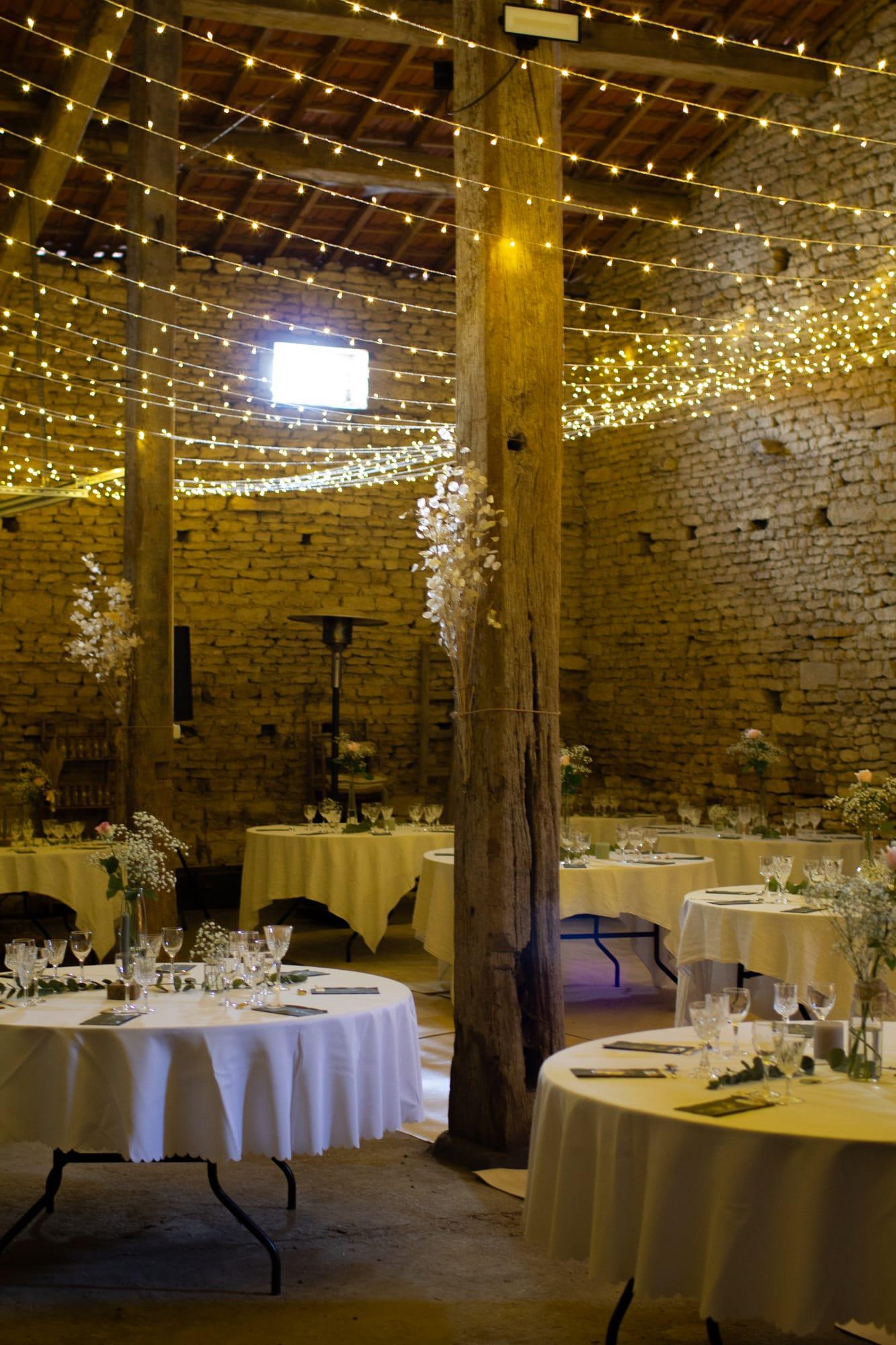 Grange en pierre illuminée de guirlandes, tables rondes nappées de blanc